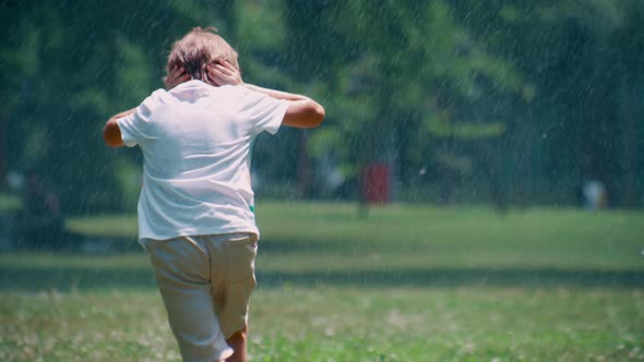 Little Boy Running Under Sprinklers in Park Hiding Ears From Water Droplet Alone