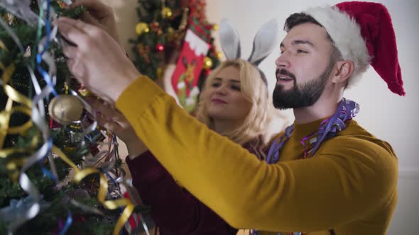 Close-up of Beautiful Caucasian Couple Hanging Christmas Decorations on New Year Tree and Admiring