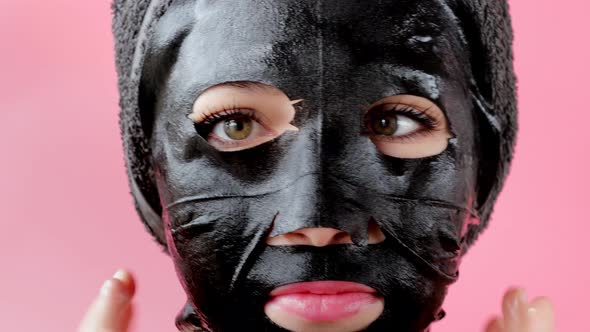 Young woman apply black cosmetic fabric facial mask on pink background