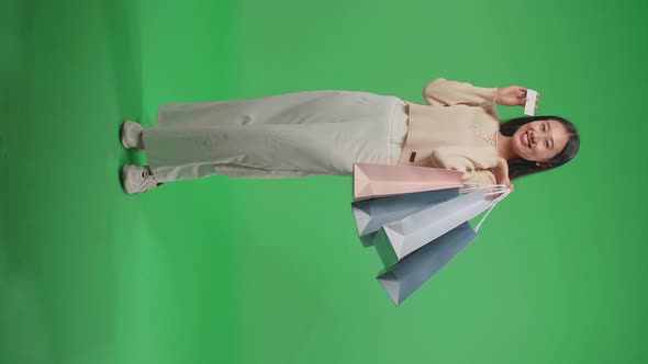 Full View Of An Asian Shopping Woman Holding A White Card And Shopping Bags On Green Screen