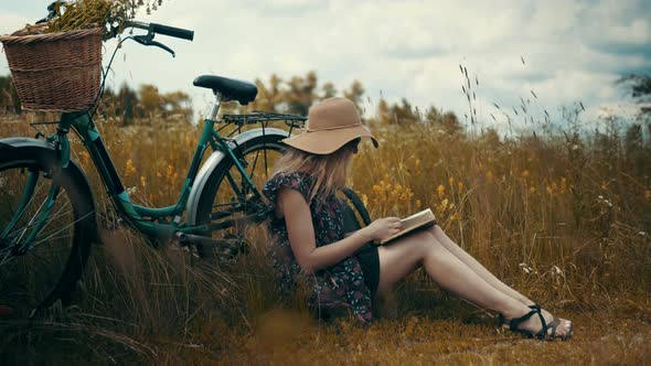 Woman Sitting On Flower Meadow And Reading Book Diary. Lady In Hat Enjoying Weekend On Holiday.