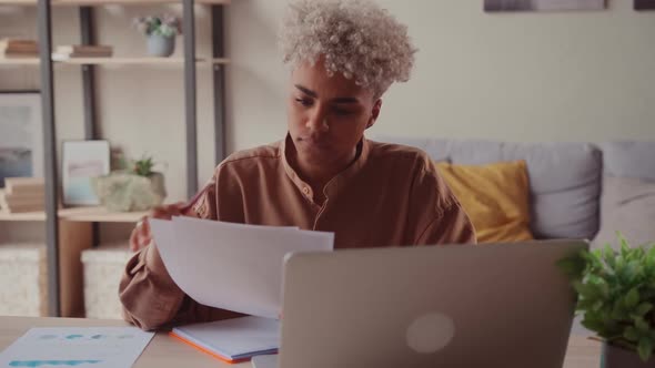 African Female at Home Table Reads Reports Compares Data on Paper and on Laptop