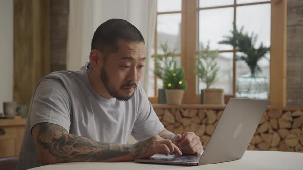 A Tottooed Asian Adult Man is Browsing on His Laptop with Various Facial Expressions