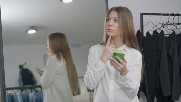 Charming Slim Woman Thinking Surfing Internet on Smartphone Standing in Retail Store Indoors
