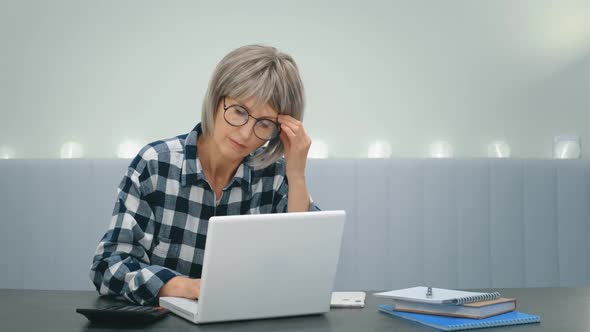 An Elderly Tired Woman with Glasses is Sitting at a Laptop and with Difficulty Overcoming Drowsiness