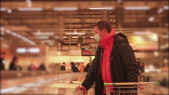 A Man with a Trolley in a Supermarket Looks at Prices with a Mask on His Face