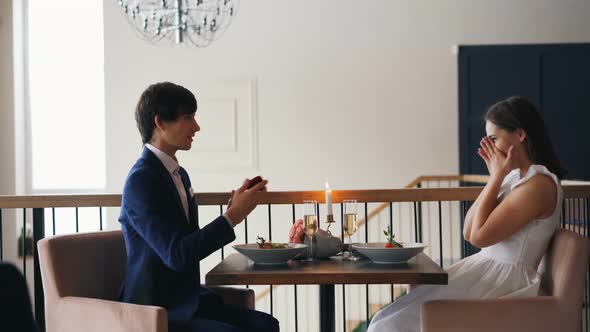 Loving Young Man Is Making Marriage Proposal To Happy Girl at Date in Restaurant Sitting at Table