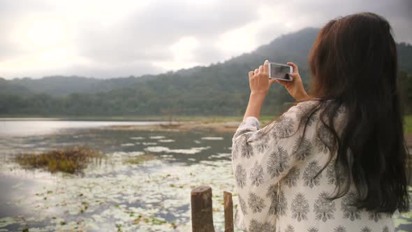 Young Mixed Race Traveler Girl Taking Photo of Beautiful Morning Lake and Mountains View Using