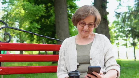 Elderly Woman with a Smartphone in Her Hands Sits on a Bench Surfing the Internet and Researching