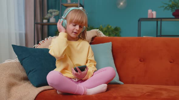 Child Girl Kid with Smartphone in Headphones Dancing While Listening Music at Home Alone on Sofa