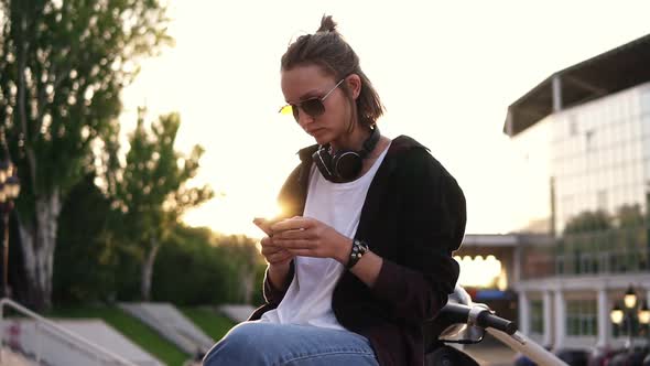 A Fashionable Modern Girl Sits on a Moped and Texting on Her Mobile Phone