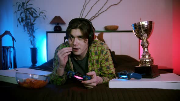 Young Man in Headphones Lying on the Bed Playing Video Game Online - Talking in the Mic