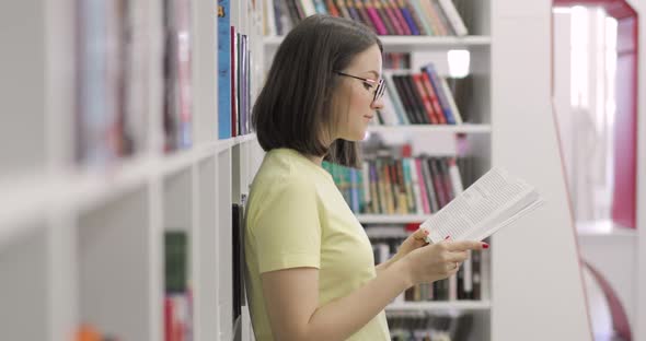 Young Beautiful European Female Student Wearing Glasses is Reading Book in College Library