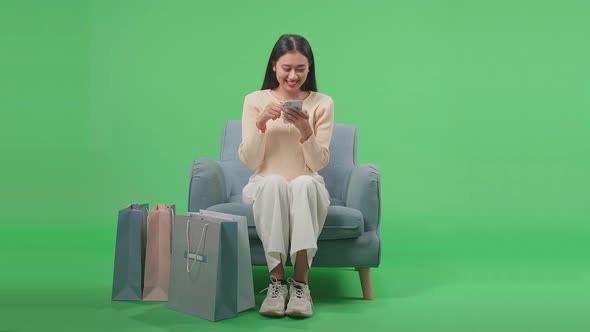 Front View Of Asian Woman Sitting On Sofa With Shopping Bags Enjoy Using Mobile Phone On Green Scree