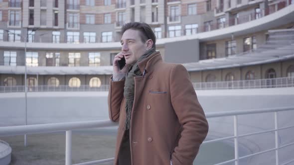 Portrait of Attractive Successful Man in Brown Coat Standing in the City Street Talking By Mobile