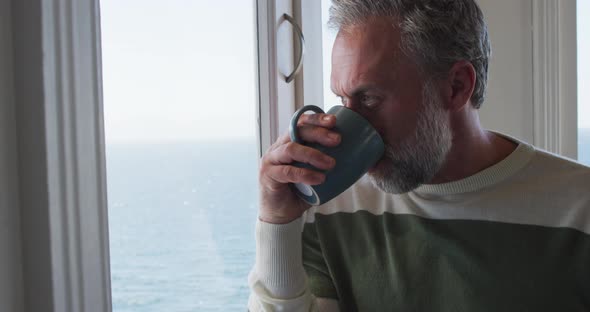 Caucasian mature man looking through the window and drinking coffee