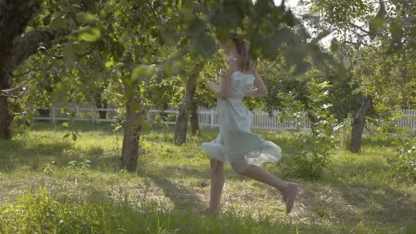 Attractive Young Woman in Straw Hat and Long White Dress Running Through the Green Summer Garden