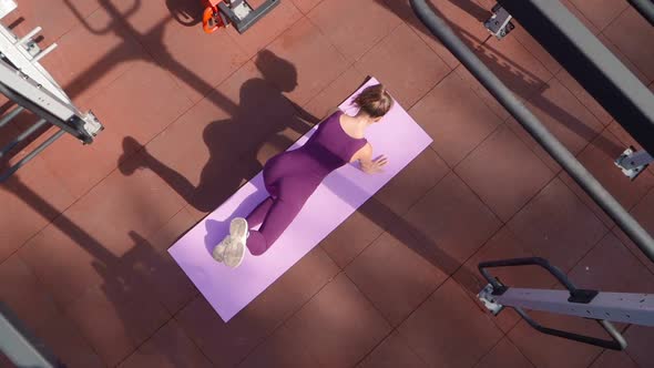 Young Athletic Woman in a Sports Purple Tracksuit Does an Exercise on the Buttocks on Workout Ground