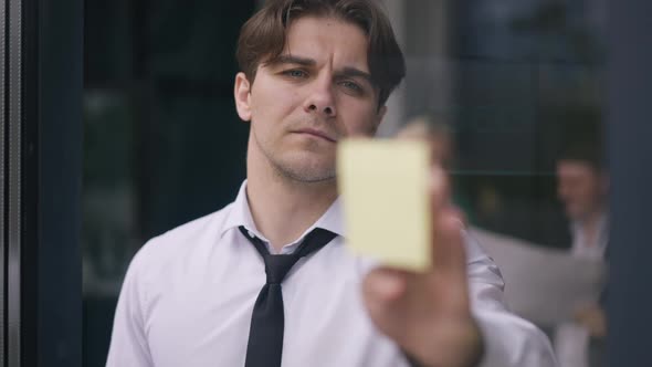 Portrait of Focused Man Sticking Adhesive Notes on Glass Door with Blurred Colleagues Reflecting