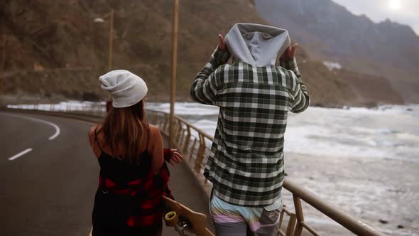 Two Young People Travel Buddies Walking Along a Highway Against a Background of Sea Coast