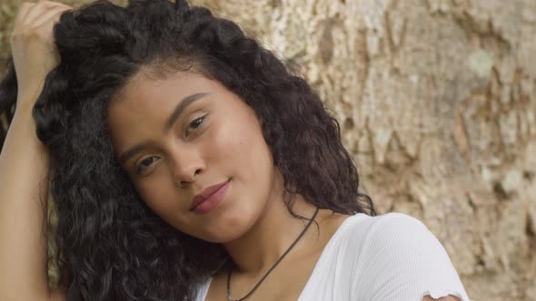 A young curly hair hispanic girl playing in her hair while standing at a large tree trunk on a sunny