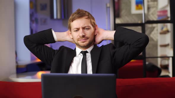 Young man in suit sitting on the couch with laptop computer lazily stretching at home office