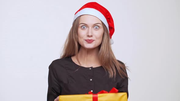 Young Beautiful Excited Fairhaired Girl in Christmas Hat Holding Gift Box Showing Thumb Up Over