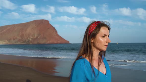 Woman in Sunglasses and Blue Dress Walking Along a Black Volcanic Beach