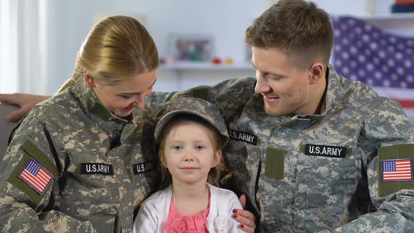 Male and Female Soldiers Hugging Daughter in Military Cap, Smiling on Camera