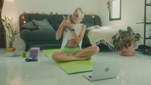 Mature Woman Sits with Cat on Yoga Mat in Front of Laptop in Preparation for Online Yoga Class