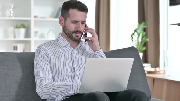 Young Businessman with Laptop Talking on Smartphone at Home