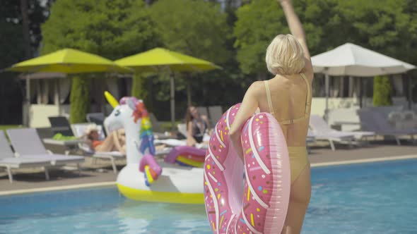 Back View of Confident Blond Woman with Donut Swimming Ring Coming To Pool and Waving To Blurred