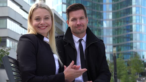 A Businessman and a Businesswoman Sit on a Bench and Applaud To the Camera in an Urban Area