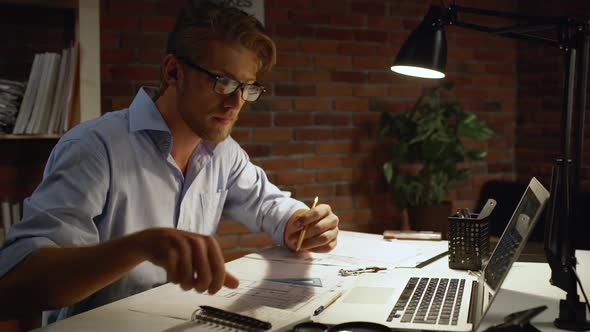 Young Successful Acrchitect with Light Beard and Glasses Sitting Drawing with Pencil with Paper and