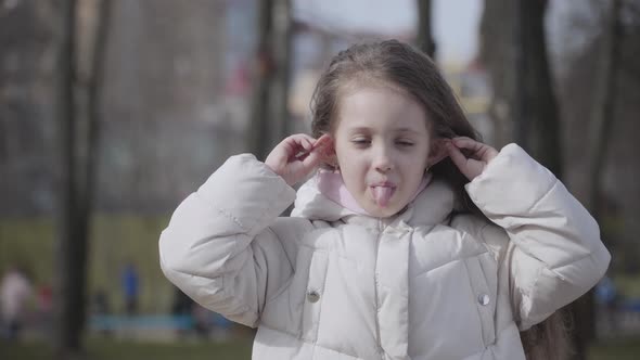 Portrait of Cute Caucasian Little Girl Pulling Ears and Showing Tongue. Funny Brunette Child with