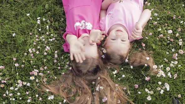 Two Cute Little Girl Lying Upside Down on Meadow Grass and Flowers Making Air Kiss and Waving Hand