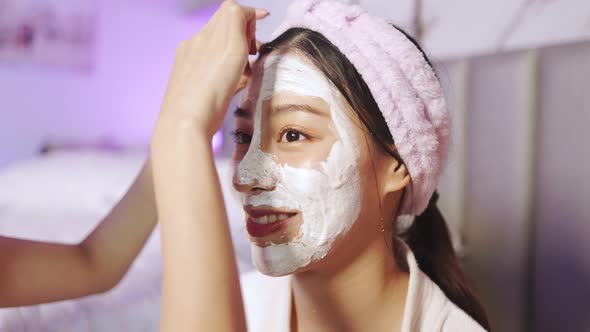 Beautiful young woman in white bathrobe applying a revitalizing  mask her friend's face.