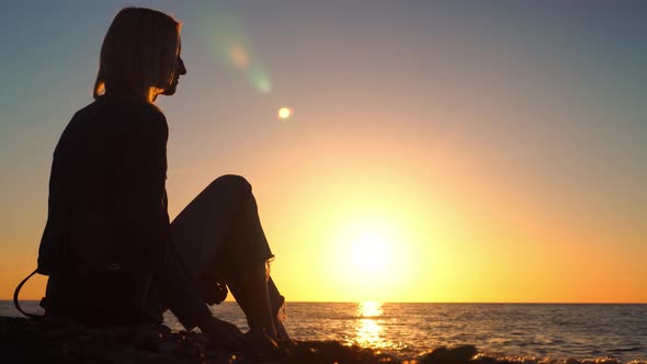 woman sitting on pebble beach by sea and throwing stones into water.