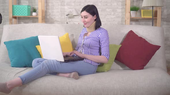 Young Woman Sits on the Couch with a Collar Around Her Neck and Uses a Laptop
