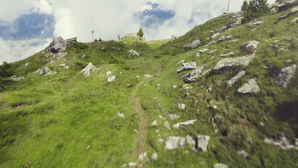 Flying along a grass covered mountain top with a cross and a small trail leading up with clouds arou