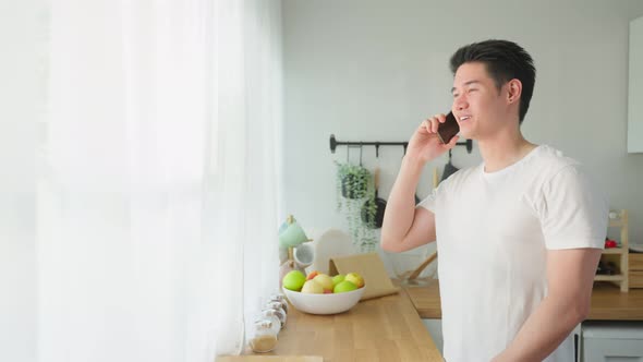 Asian handsome man talking on mobile phone call stand in kitchen at home.