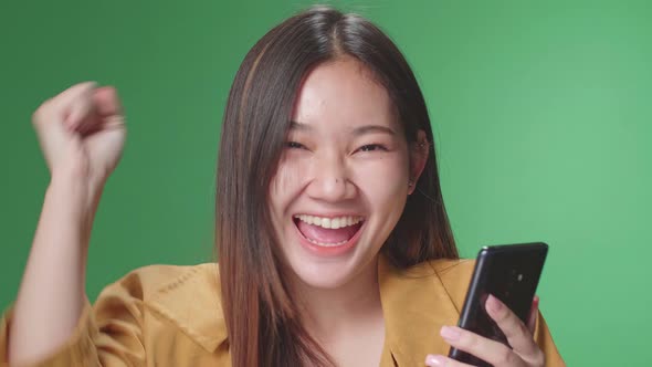 Young Asian Woman Celebrating With Mobile Phone While Standing On Green Screen In The Studio