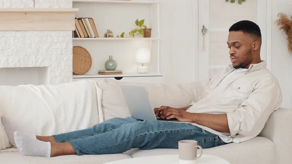 Focused African Guy Student Lying on Couch with Laptop Typing Browsing on Web Study Remotely From