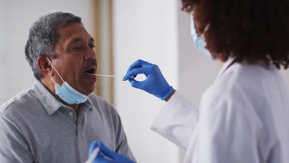 Mixed race female doctor wearing mask doing swab test on senior man at home