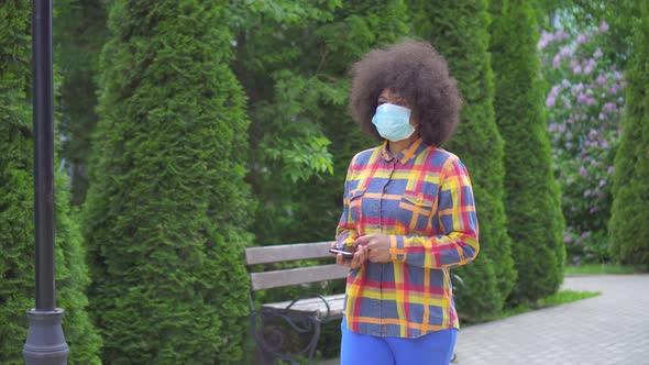 Black American Woman with an Afro Hairstyle in a Protective Medical Mask with a Smartphone in His