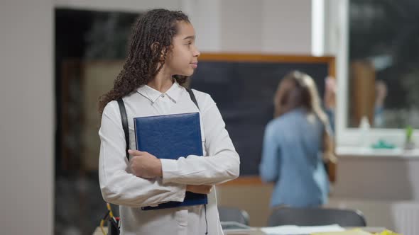 Middle Shot Portrait of Positive Cute African American Schoolgirl with Workbook Posing in Classroom