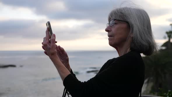An old lady on vacation happy and smiling while taking a selfie with her smart phone on the ocean in