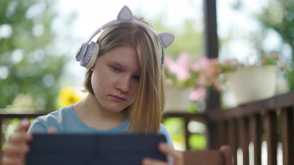 Teenage Girl Sits At Wooden Table In a Summer Cafe In Headphones With Phone