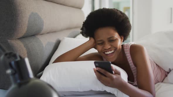 Happy african american woman laying in bed, using smartphone in bedroom