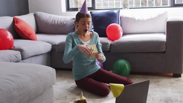 Surprised woman in party hat holding a gift box while having a video call on laptop at home